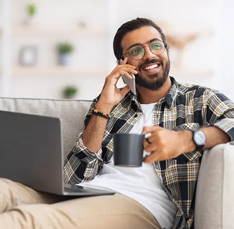 Man with glasses on having a phone call, smiling, holding a cup, and has the laptop on his lap.